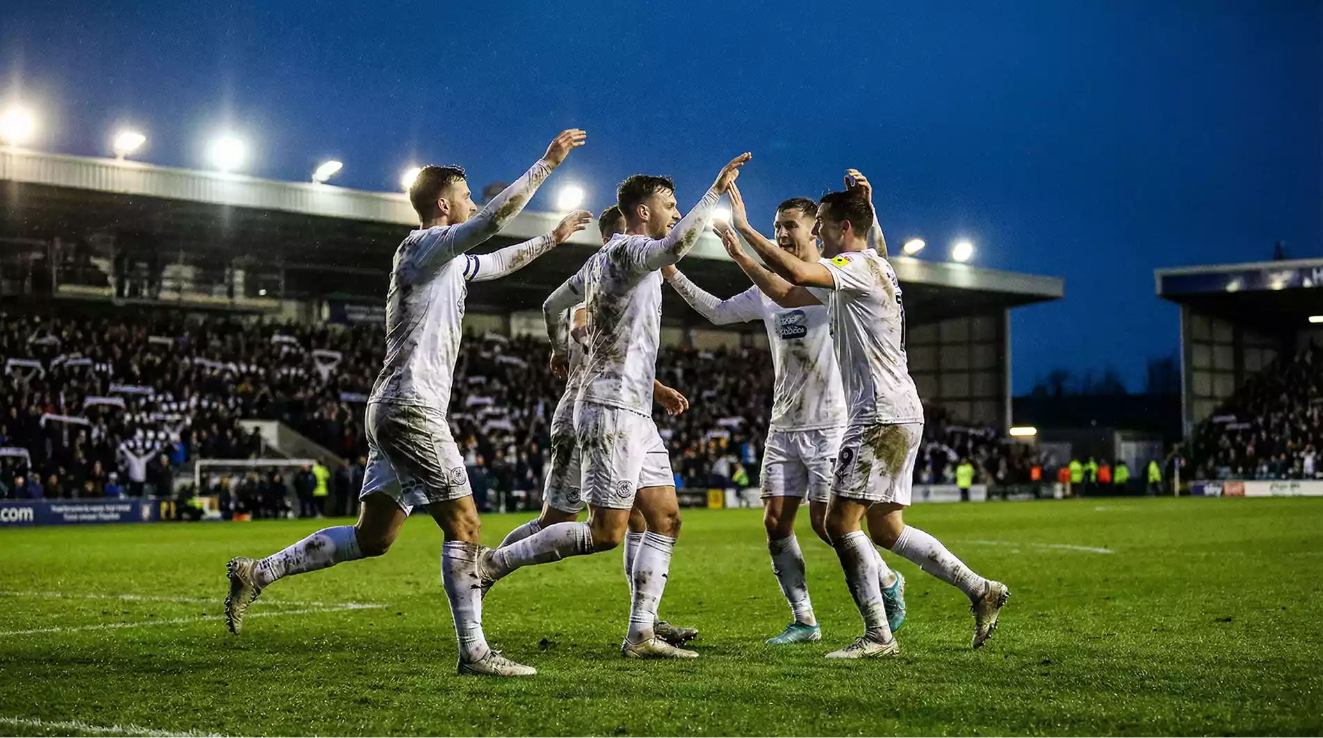 Football players in white kits celebrating a goal together on a lush green pitch under stadium floodlights at night
