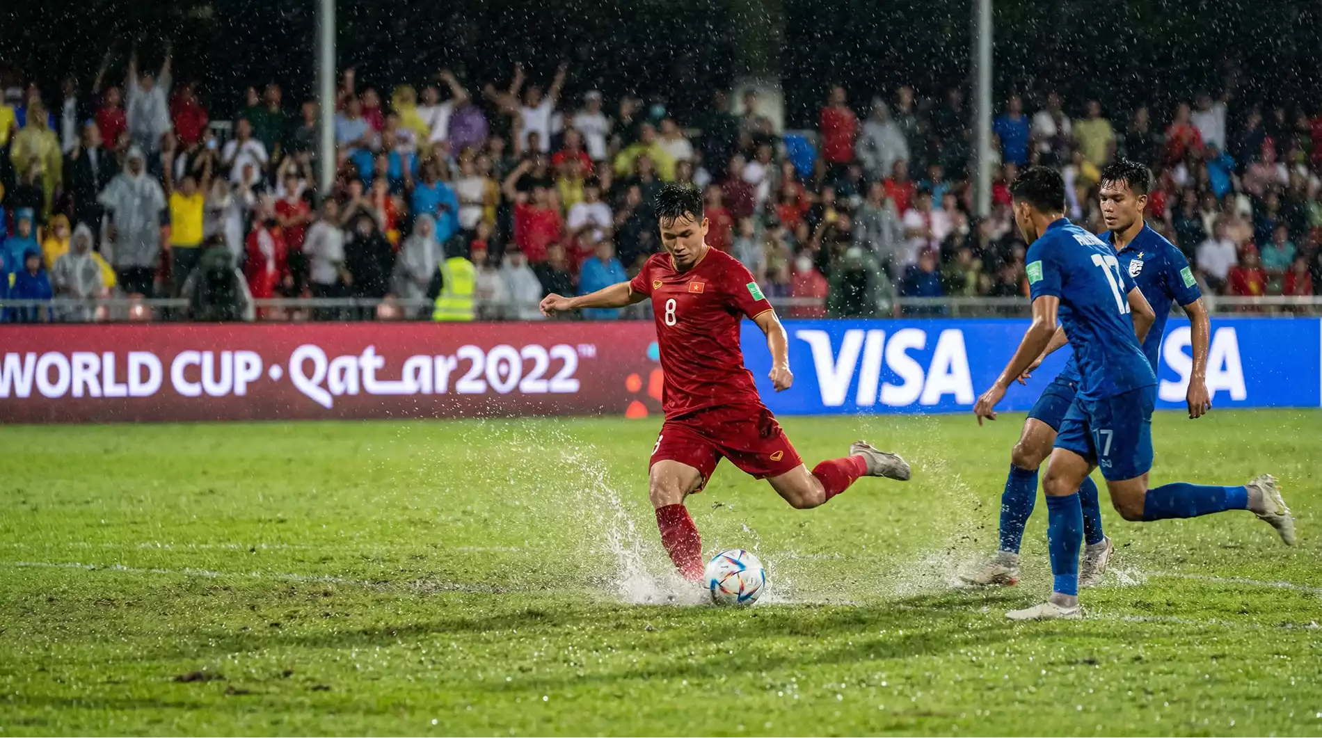 Football midfielder in a red kit dribbling past two defenders on a rain-soaked World Cup pitch with a roaring crowd in the background