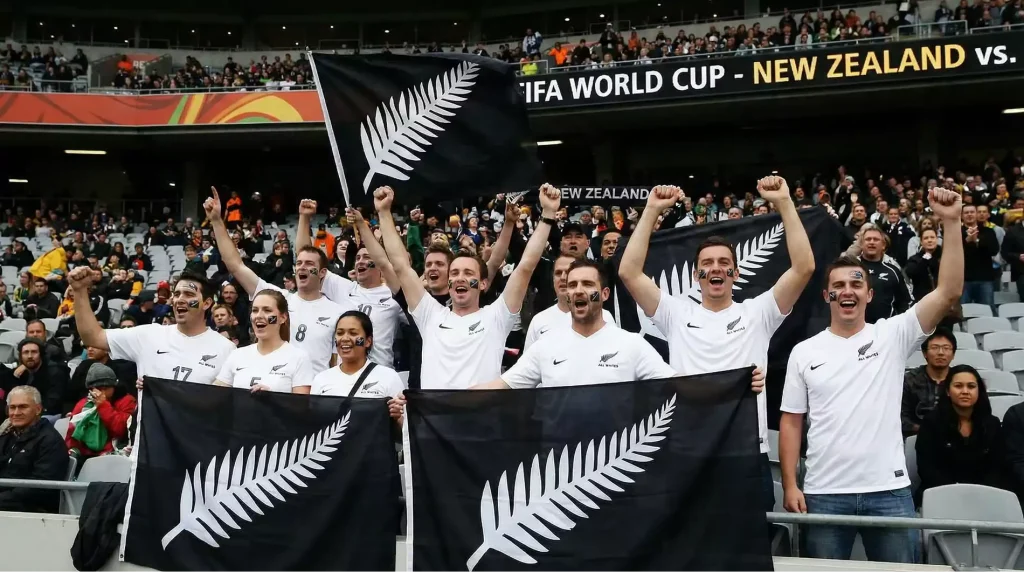 New Zealand supporters in All Whites jerseys celebrating in stadium stands with silver fern flags