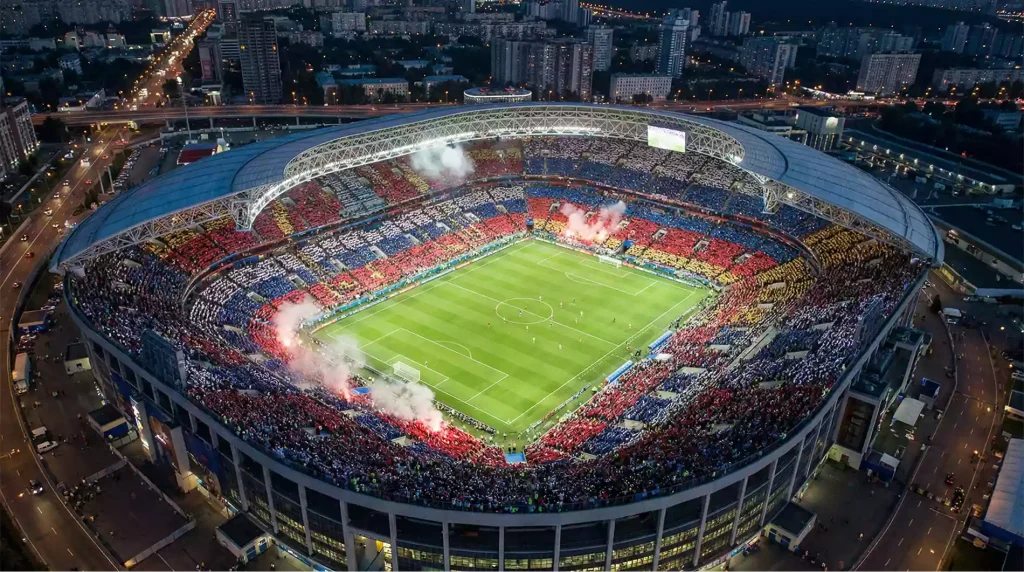 Aerial view of World Cup stadium at night with 80,000 fans creating flag mosaic in the stands
